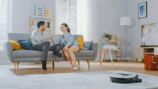 Robô inteligente aspirador de pó sugando o pó de um tapete. Casal bonito está sentado em um sofá e falando no fundo. Dispositivo de eletrodomésticos tecnológicos move-os para além deles. — Fotografia de Stock