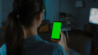 Genç Yakışıklı Kadın Akıllı Telefonunu Chroma Key ile Tutuyor. Model için harika bir görüntü. Işıkları Kapalı Oturma Odasında Sıcak Bir Gece. Teknolojiden etkilenmiş..
