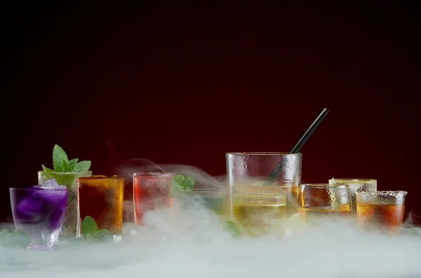 Various alcoholic cocktails with ice and mint framed by smoke on a red background — Stock Photo, Image