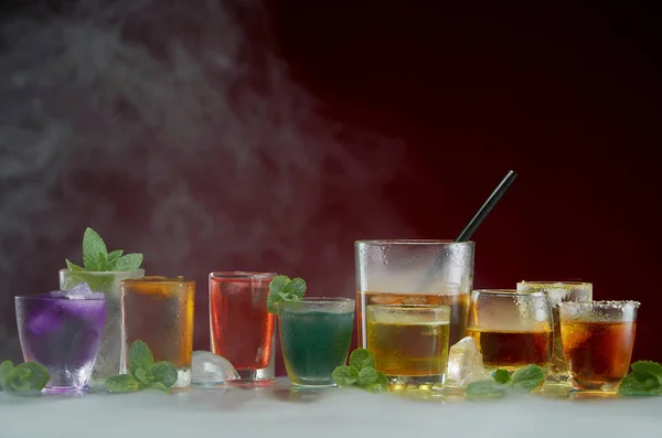 Various alcoholic cocktails with ice and mint framed by smoke on a red background — Stock Photo, Image