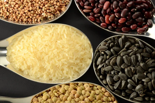 Rice, buckwheat, seeds, lentils, beans in spoons on a dark background.