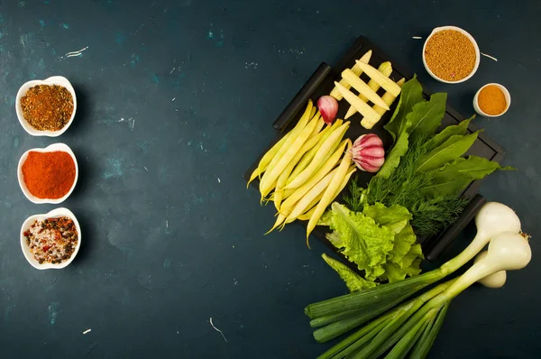 LAS VEGETALES EN LA CAJA DE LA PIEDRA UNA OSCURIDAD ANTECEDENTES. JOVEN GREENS ONION GARLIC ZUCCHINI BRIGHT SPICES ESTAR EN UNA bandeja de madera con las manos en una OSCURIDAD TEXTUADA ANTECEDENTES. — Foto de Stock