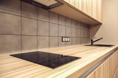 Gri lavabosu ve su musluğu olan kare fayanslı mutfak önlüğü prize yakın ahşap bir tezgah, dolap ve siyah elektrikli hob