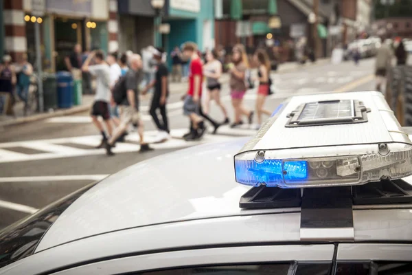 A bue light flashes on the top of a police cruiser as pedestrians cross the street in the background.