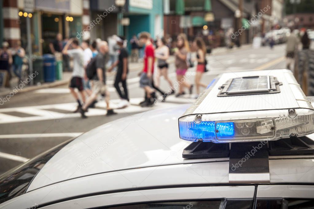 A bue light flashes on the top of a police cruiser as pedestrians cross the street in the background.