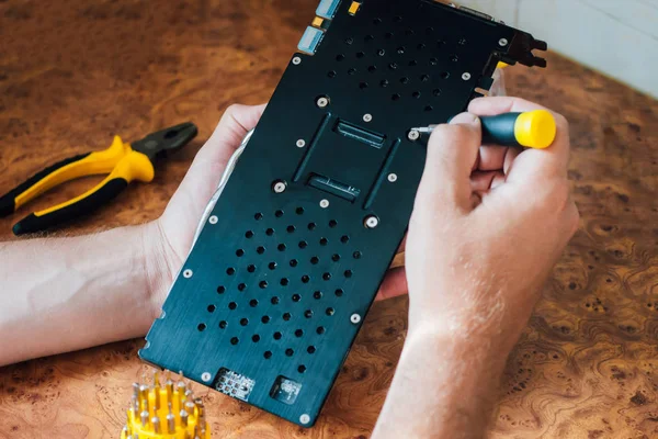 Computer video card repair. Male hands with a yellow screwdriver on a wooden background