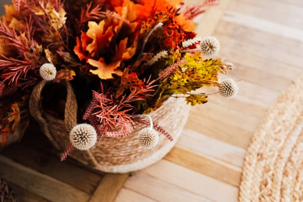 Sonbahar yaprakları ve kurumuş çiçeklerden oluşan renkli bir buket dokuma sepetinde sergilenir. Düzenlemede turuncu kırmızı ve sarı ılık renkler sergileniyor. Dekor rahat bir atmosferi vurguluyor..