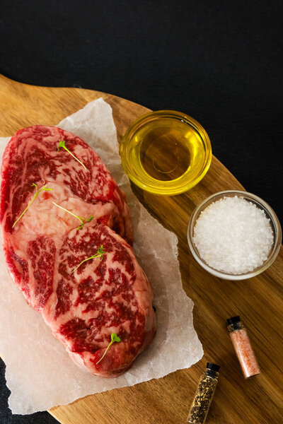 Two marbled beef steaks are placed on parchment paper atop a wooden cutting board. Olive oil sea salt and assorted spices are arranged nearby. Ingredients suggest preparation for grilling.
