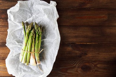 Bir demet taze yeşil kuşkonmaz düzgünce bağlanmış ve buruşmuş parşömen kağıdının üzerinde duruyor. Kırsal ahşap masa bu canlı sebze gösterisine sıcaklık ve cazibe katıyor..