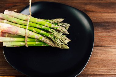 Bir demet taze yeşil kuşkonmaz sicimle güzelce bağlanır ve parlak siyah bir tabağa yerleştirilir. Kırsal ahşap arka plan sağlıklı bir yemek için mükemmel sebzelerin doğal güzelliğini arttırır..