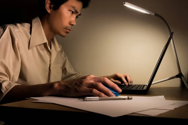 A busy business man is sitting on the chair for working overtime at office at night. He use a led lamp for lighting his desk for writing and typing laptop for research information.