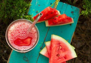 Metal bir pipetle bardakta taze karpuz suyu manzarası. Turkuaz ahşap bir yüzeyde karpuz dilimleri