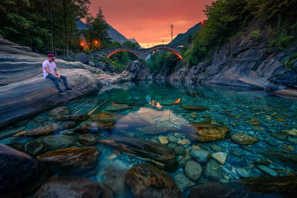 O turista desfruta do pôr do sol em um rio perto da ponte de pedra em Lavertezzo, Suíça — Fotografia de Stock