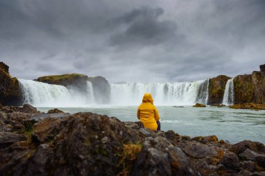 Sarı ceketli turist İzlanda 'daki Godafoss şelalesinde dinleniyor.