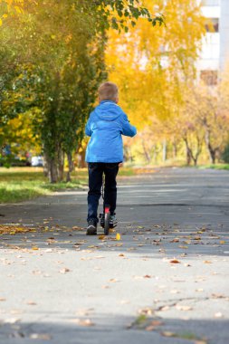Mavi ceketli bir çocuk sonbaharda şehir caddesinde scooter sürüyor.
