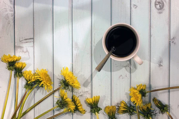 A bouquet of yellow dandelions on a sky-colored tree background. White Cup of coffee. Coffee and flowers. Still life of flowers on a plank table.