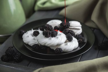 Siyah bir tabakta beyaz krema ve marşmelov, kırmızı şurupla doldurulmuş. Çilek şurubu. Tatlı böğürtlenlerle süslenir. Siyah böğürtlen ve beyaz kurabiye. Karanlık ahşap bir masada hayat.