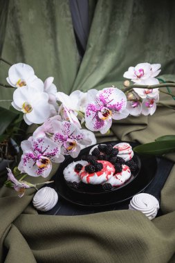 Siyah bir tabakta beyaz krema ve marşmelov, kırmızı şurupla doldurulmuş. Çiçeklerle ve pişirmeyle dolu bir hayat. Hafif Orkide. Çilek şurubu. Tatlı böğürtlenlerle süslenir. Siyah böğürtlen ve beyaz kurabiye. Karanlık ahşap bir masada hayat.