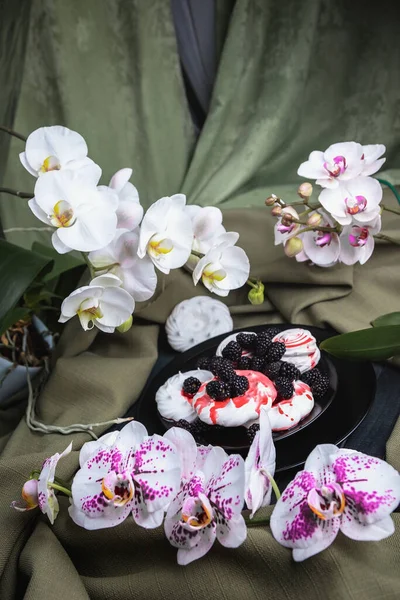 Still life of flowers Orchid and dessert. Tender, delicious marshmallows and meringue, on a black plate.