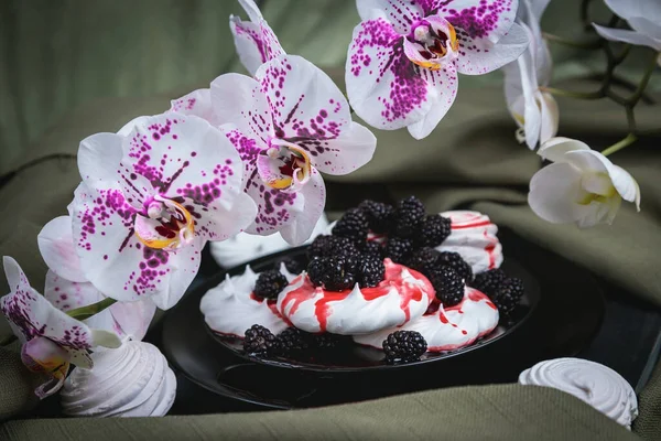 Still life of flowers Orchid and dessert. Tender, delicious marshmallows and meringue, on a black plate.