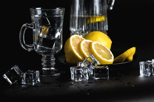 Still life on a black background. Glass and ice. Cold drinking water. Yellow lemon in a black plate.