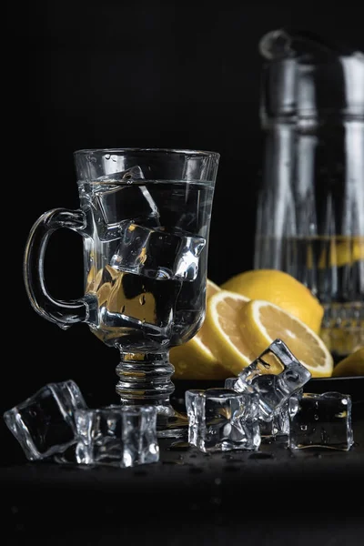 Sliced lemon on a black plate. A decanter and a tall glass of water and ice. Still life of glass objects on a black background. Cold lemonade. Yellow on black.