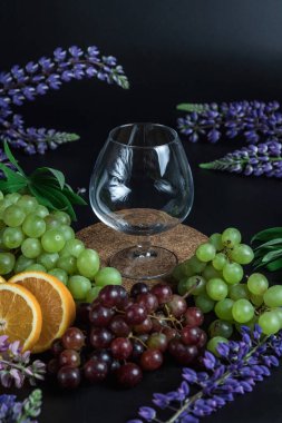 Üzüm ve portakallar arasında konyak bardağıyla sakin bir hayat. Siyah arka planda hala hayat var. Çiçekler lupinli..