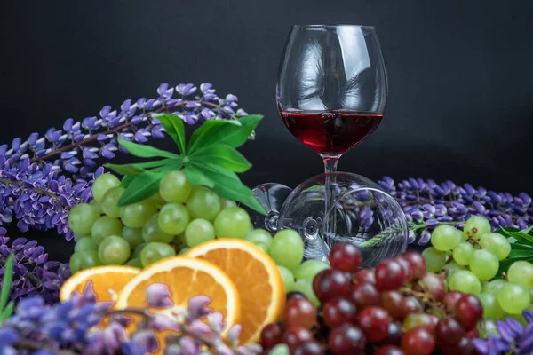A glass of red wine is on the table. Beautiful still life on a black background. Still life of grapes, oranges and lupine flowers.