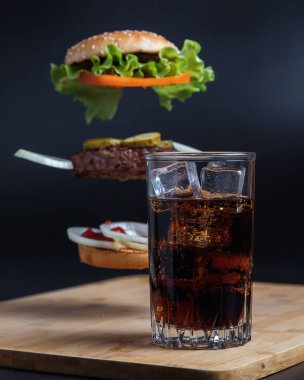 Klasik Burger. Hamburger ve kola dolu bir hayat. Buzlu uzun bir bardakta iç. Hamburgerin bileşenleri havada siyah bir zemin üzerinde uçuyor. Et pirzolası, soğan, domates, marul ve peynir..