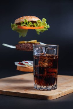Klasik Burger. Hamburger ve kola dolu bir hayat. Buzlu uzun bir bardakta iç. Hamburgerin bileşenleri havada siyah bir zemin üzerinde uçuyor. Et pirzolası, soğan, domates, marul ve peynir..