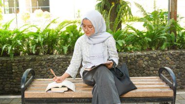 Asyalı tesettürcü kız kampüs bahçesinde notlar yazıyor.