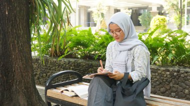 Asyalı tesettürcü kız kampüs bahçesinde notlar yazıyor.