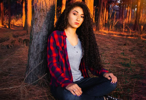 Black teen girl with voluminous curly hair, sitting in the forest at sunset, leaning against a tree, wearing a plaid shirt. Afro beauty, black beauty. Autumn weather