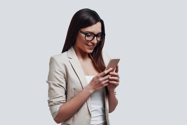 Business chat. Beautiful young woman looking at smart phone while standing against grey background