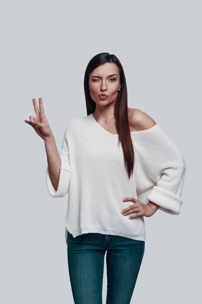 Símbolo de paz. Atractiva joven mujer mirando a la cámara y haciendo gestos mientras está de pie sobre un fondo gris —  Fotos de Stock