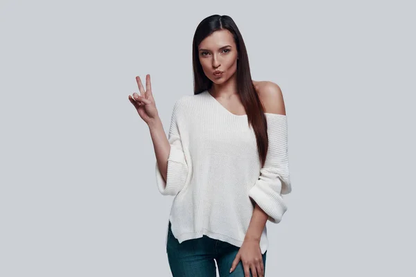 Paz. Atractiva joven mujer mirando a la cámara y haciendo gestos mientras está de pie sobre un fondo gris — Foto de Stock