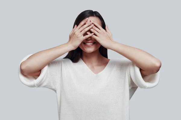 See nothing. Attractive young woman covering eyes with hands and smiling while standing against grey background — Stock Photo, Image