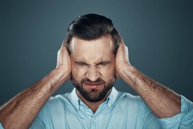 Sorun. Gri arka planda ayakta iken elleri ile kulakları kapsayan sinirli genç adam