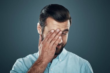 Yorgun hissediyorum. Yakışıklı genç adam el ile göz kapsayan ve gri arka planda ayakta iken gülümsüyor