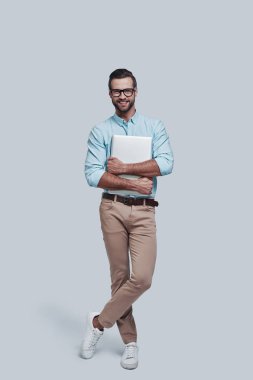 Genç ve başarılı. Tam uzunluğu iyi görünümlü genç adam laptop taşıyan ve gri arka planda ayakta iken kamera bakıyor