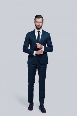 Onun tarzında emin. Kamera bakıyor ve gri arka planda ayakta iken onun kol ayarlama yakışıklı genç adamın tam uzunluğu