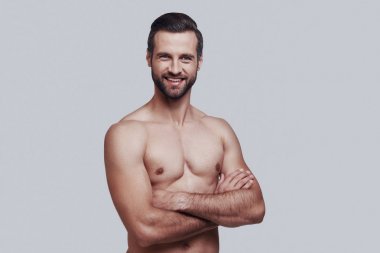 Kendine güvenen ve yakışıklı. Yakışıklı genç adam kamera bakıyor ve gri arka planda ayakta iken gülümsüyor