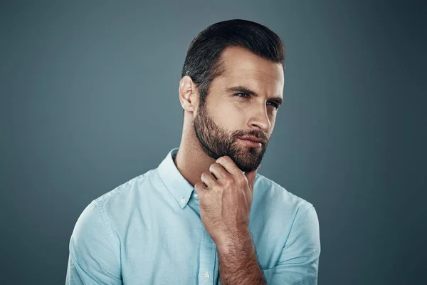 Mirando hacia el futuro. Joven guapo mirando hacia otro lado y manteniendo las manos en la barbilla mientras está de pie sobre un fondo gris — Foto de Stock