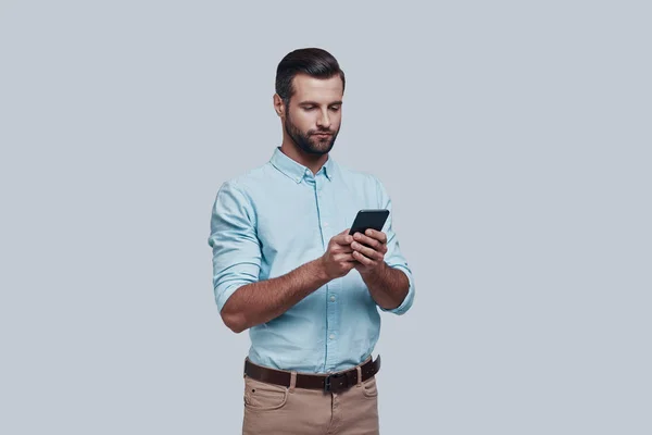 중요 한 비즈니스 메시지입니다. 회색 배경에 서 있는 동안 잘생긴 젊은 남자 스마트 폰을 사용 하 여 — 스톡 사진