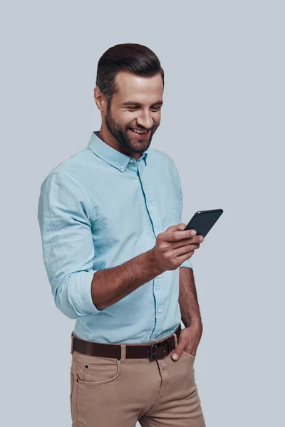 Ontvangen van goede feedback. Knappe jonge man met behulp van slimme telefoon en glimlachen, terwijl staande tegen grijze achtergrond — Stockfoto