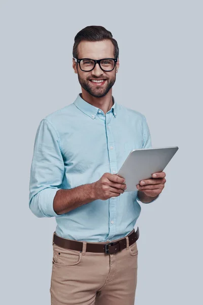 Klaar om te helpen. Knappe jonge man met behulp van digitale Tablet en glimlachen, terwijl staande tegen grijze achtergrond — Stockfoto