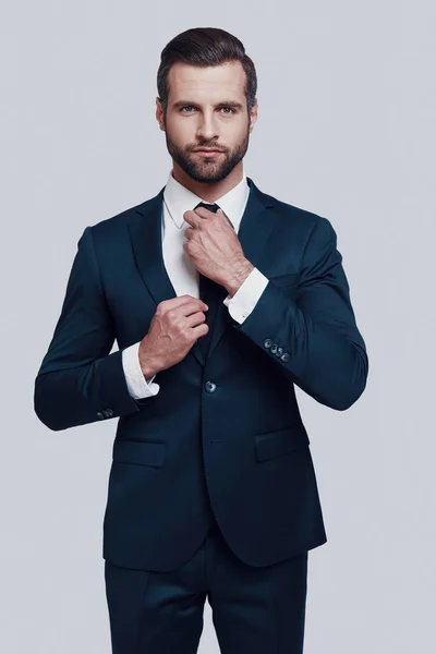Good looking entrepreneur. Handsome young man looking at camera and adjusting his necktie while standing against grey background — Stock Photo, Image