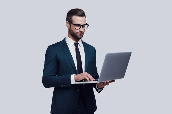 Prestare attenzione ad ogni dettaglio. Bello giovane uomo che utilizza il computer portatile e sorridente mentre in piedi su sfondo grigio — Foto Stock