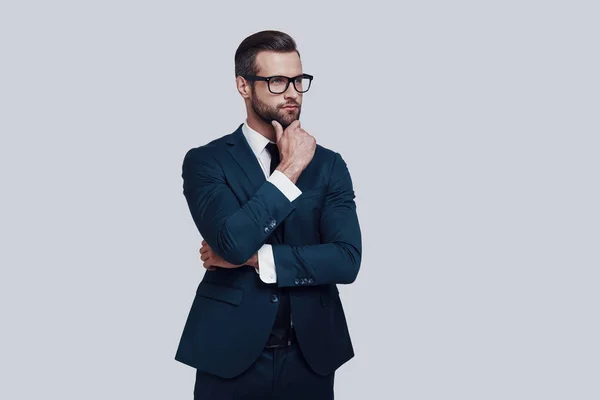 Thinking about... Handsome young man keeping hand on chin and looking away while standing against grey background — Stock Photo, Image