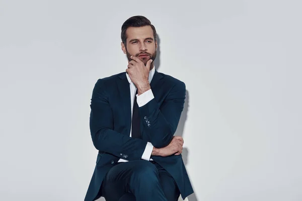 Hard choice. Handsome young man looking away and keeping hand on chin while sitting against grey background — Stock Photo, Image